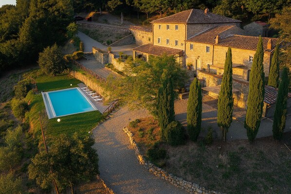A large stone building with a swimming pool, surrounded by trees and a gravel path.