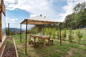 Una mesa y banco de madera bajo una zona sombreada, rodeados de vegetación y vistas al paisaje.