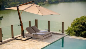 A poolside lounge area with a cushioned seating arrangement, a large umbrella, and a glass balustrade overlooking a body of water. - Tabebuia Spa & Safari Resort (Fort Portal)