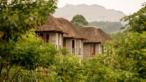 A rustic building with thatched roofs, surrounded by lush greenery and hills in the background. - Tabebuia Spa & Safari Resort (Fort Portal)