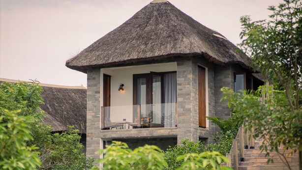 A thatched-roof bungalow with a stone exterior, a balcony with seating, and lush greenery surrounding it. - Tabebuia Spa & Safari Resort (Fort Portal)