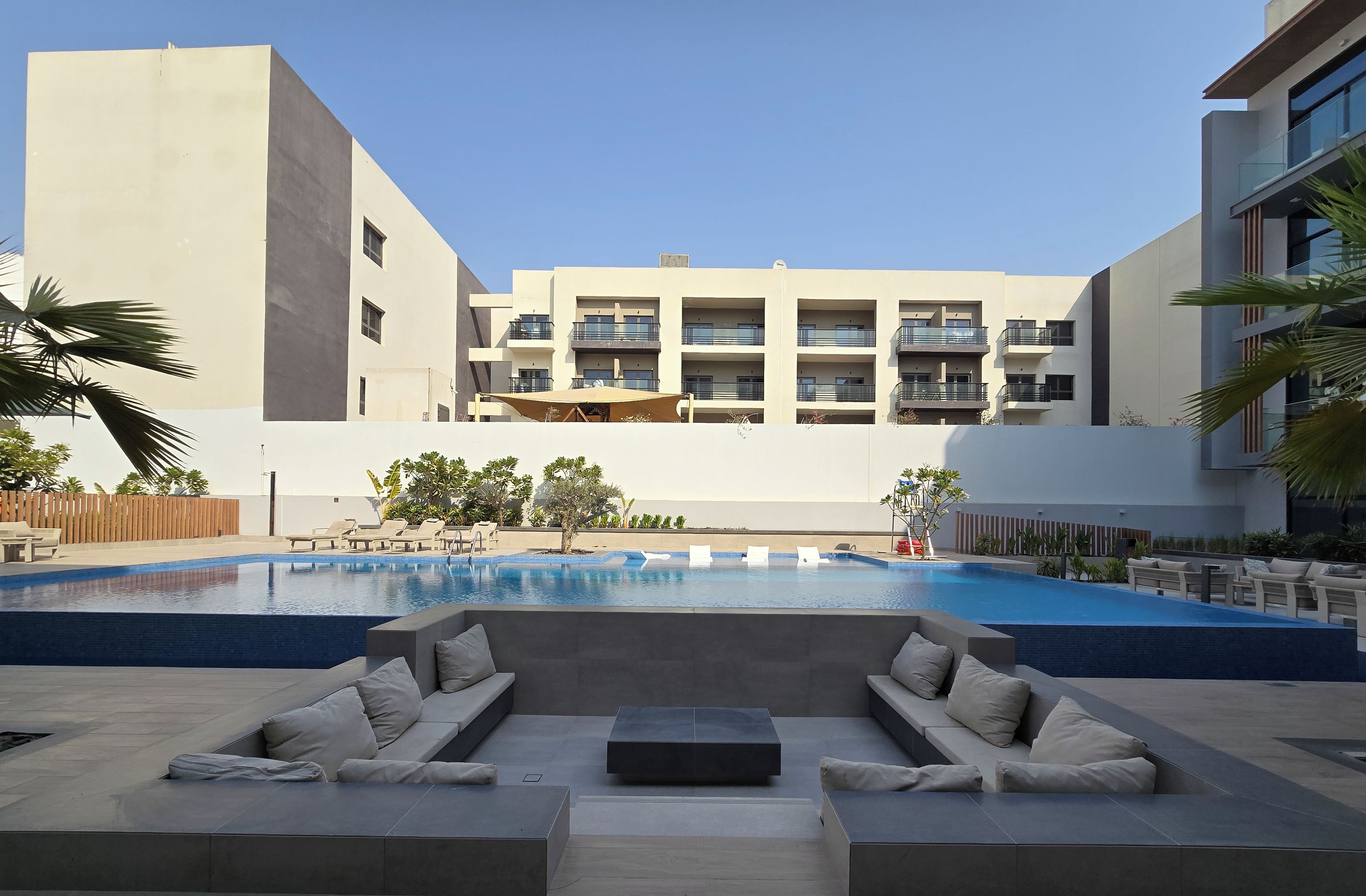 Un área de piscina exterior moderna con sillones de descanso y una zona de estar, rodeada por un edificio con balcones.