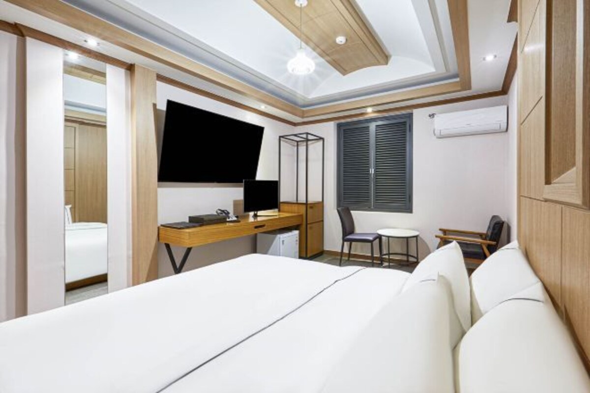 Una habitación de hotel moderna con una cama grande, un escritorio con computadora, una silla, un televisor y una ventana con persianas.