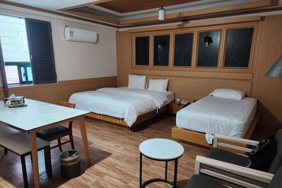 Habitación de hotel con dos camas, una mesita, una silla, un taburete redondo y una ventana con persianas.
