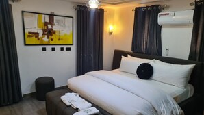 A modern hotel room with a neatly made bed, a small bedside table, a wall-mounted air conditioner, and a painting on the wall. - Blooming Apartments (Jimeta)