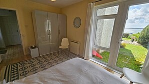 Un dormitorio con cama, armario, silla, radiador y un ventanal con vista a un jardín y al agua.