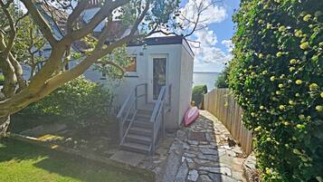 Una escalera de piedra que conduce a un edificio blanco con un pequeño ventanal, rodeado de vegetación y una valla de madera.