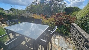 Mesa con cubierta de vidrio y cuatro sillas, ubicada en un patio de piedra con pared de piedra y vegetación exuberante al fondo.