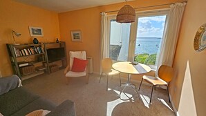 Ein gemütliches Wohnzimmer mit einer Couch, einem kleinen Tisch und Stühlen. Ein Bücherregal mit Büchern und ein Fernseher sind ebenfalls vorhanden. Ein Fenster bietet Blick auf das Meer.