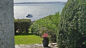 Ein gepflasterter Weg führt zu einer grünen Fläche mit Blick auf das Meer und einem Segelboot in der Ferne.