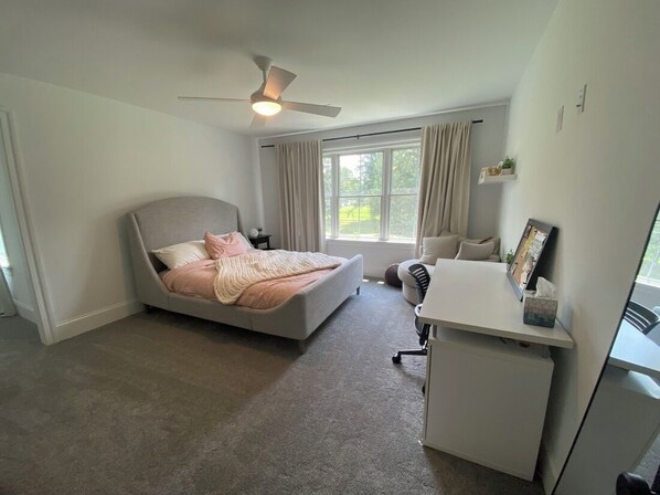 A bedroom with a bed, a desk, a chair, a ceiling fan, and a window with curtains.