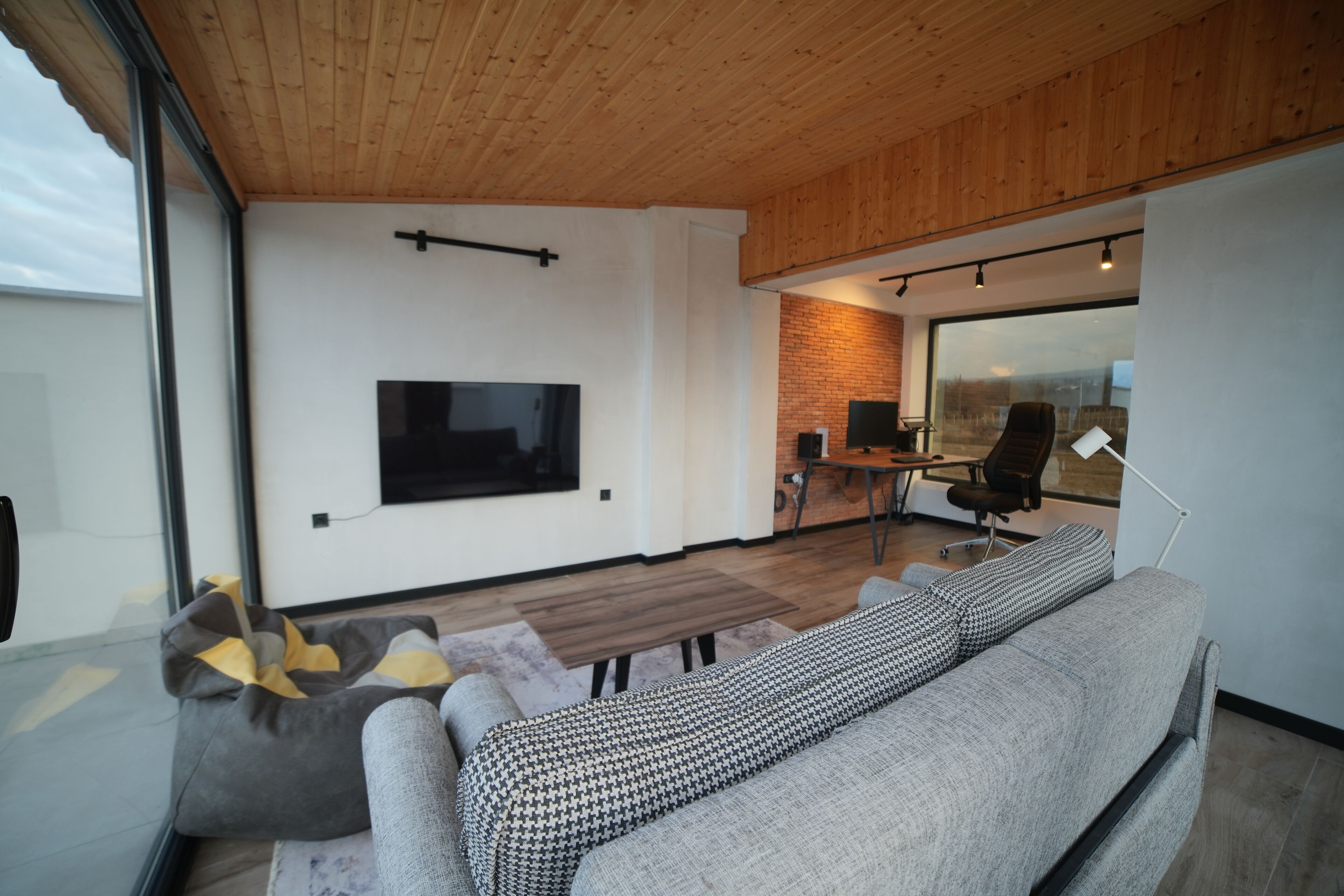 Uma sala de estar moderna com um sofá cinza, uma manta xadrez, uma mesa de centro de vidro e uma TV fixada na parede.