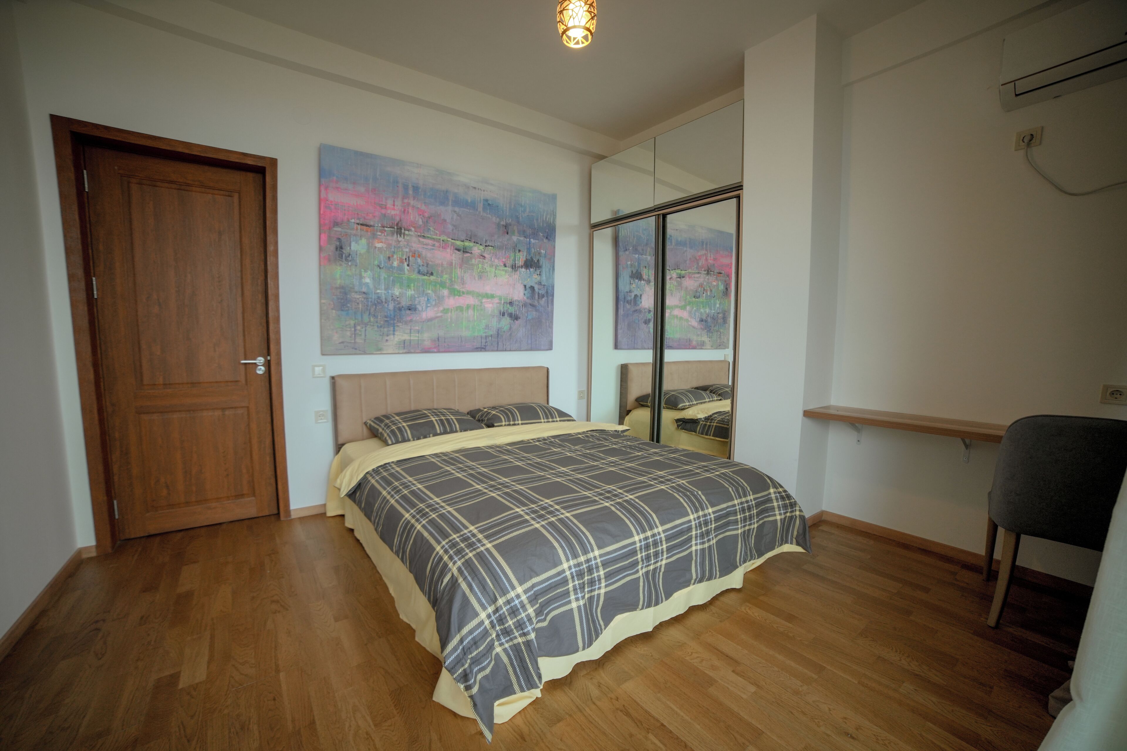 A bedroom with a wooden floor, a bed with a plaid bedspread, a wooden door, and a large mirror on the wall.
