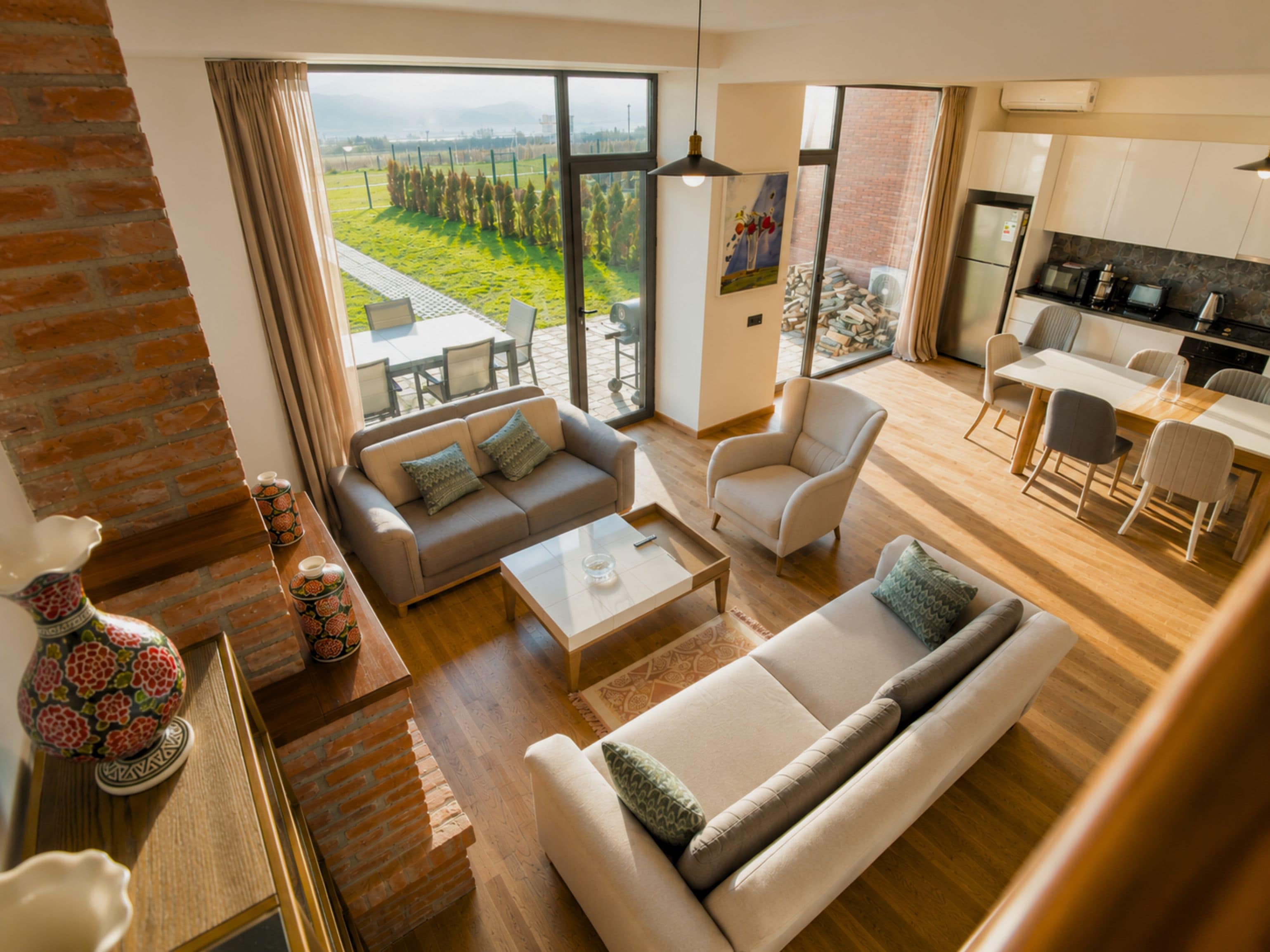 Un amplio salón con sofá, sillón y mesa de centro, una cocina con electrodomésticos modernos y un gran ventanal con vistas a un paisaje verde.