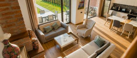 Ein großzügiger Wohnbereich mit Sofa, Sesseln und Couchtisch, eine Küche mit modernen Geräten und ein großes Fenster mit Blick auf eine grüne Landschaft.