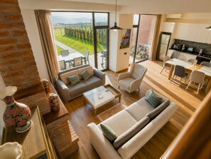 A spacious living area with a sofa, armchair, and coffee table, a kitchen with modern appliances, and a large window offering a view of a green landscape. - Matilda Signature Villas (Telavi)