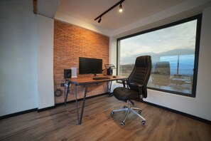 A modern office space with a brick wall, a large window, a desk with a computer, and a black office chair. - Matilda Signature Villas (Telavi)