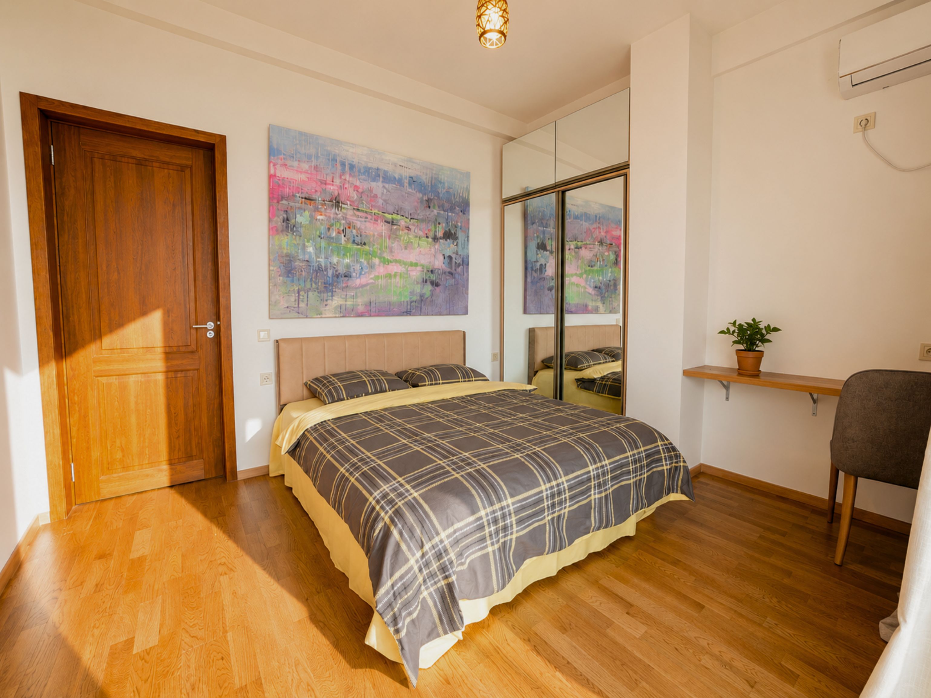 A bedroom with a bed, a wooden door, a painting, a mirror, a potted plant, and a desk with a chair.