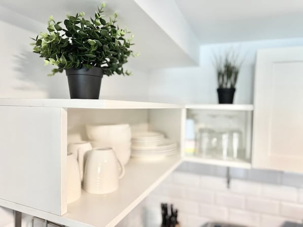 A white kitchen shelf with a potted plant, white plates, and a white mug.
