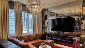 A modern living room with a leather sofa, a dining table with placemats, a large flat-screen TV, a fireplace, and a chandelier. - Lux Nash Escape•11 Beds•Hot Tub•Game Room + Pool+ Pickleball (Nashville)