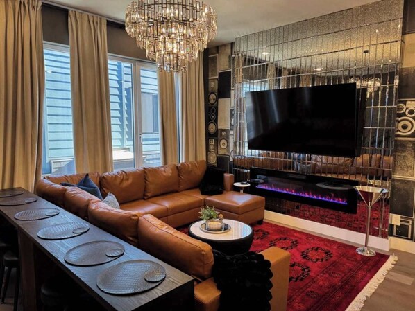 A modern living room with a leather sofa, a dining table with placemats, a large flat-screen TV, a fireplace, and a chandelier.