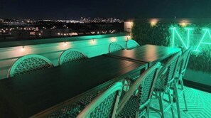 Terraço no telhado com mesa e cadeiras, um letreiro neon e vista para o horizonte da cidade.
