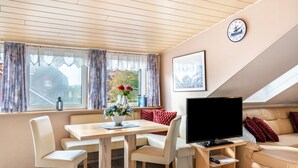 Een gezellige kamer met houten plafond, een televisie, een bank, een eettafel met stoelen en een raam met gordijnen.