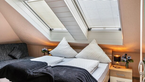 Een slaapkamer met een schuine kap, dakramen, een bed met wit beddengoed, een nachtkastje met een lamp en een bijzettafeltje met een bloevas.