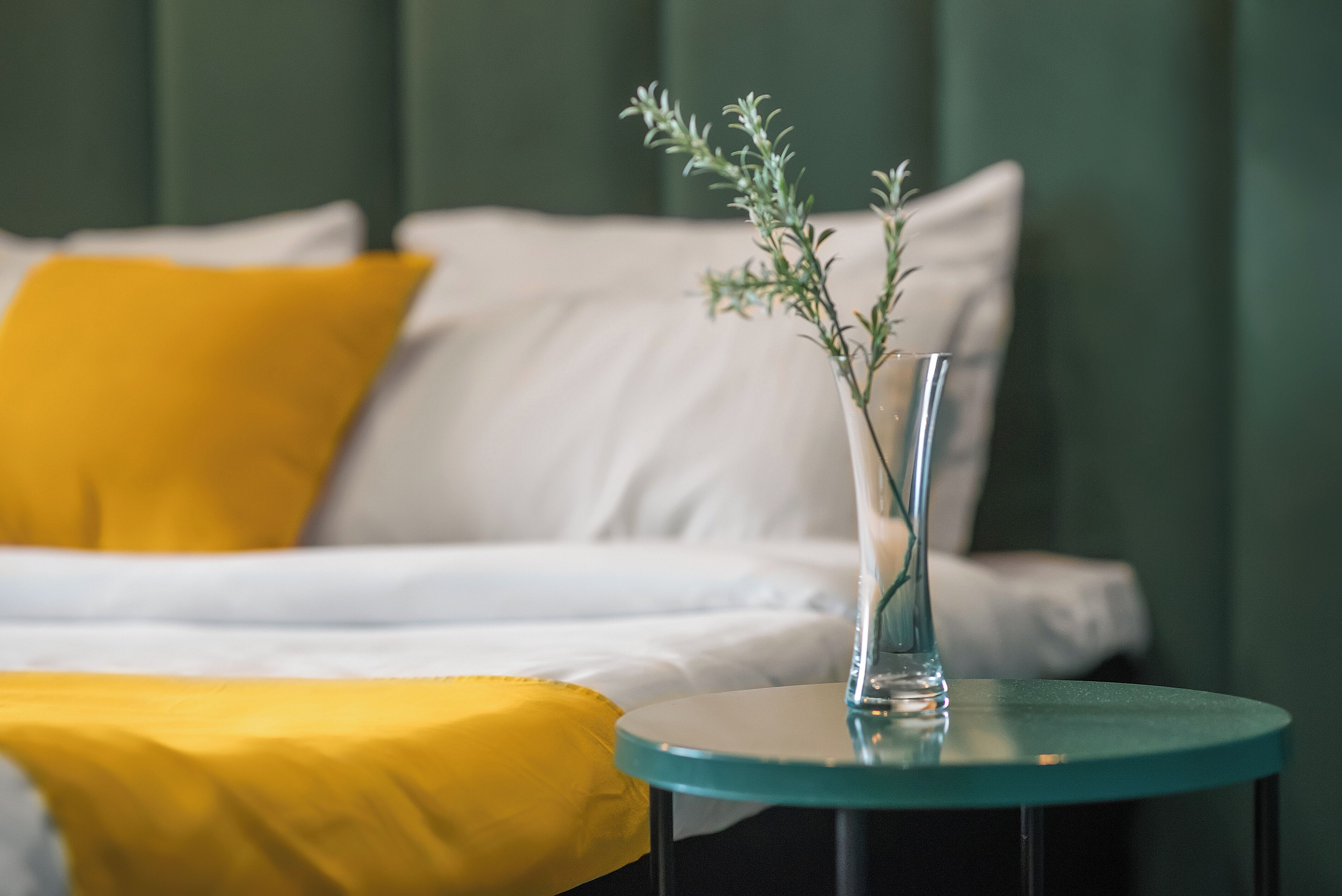 A bed with white and yellow bedding, a glass vase with a plant, and a round table beside the bed.