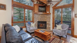 A cozy living room with a stone fireplace, leather sofa, and large windows offering a view of trees.