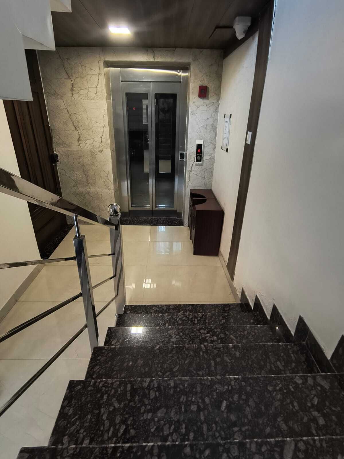 Un ascenseur moderne aux finitions métalliques, un mur en marbre et une petite poubelle placée à côté de l’escalier.