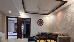 A modern living room with a sofa, chairs, a ceiling fan, and a decorative wall piece. - HomiStay (Panchkula)