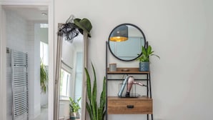 Une salle de bain équipée d’un porte-serviettes, d’un miroir, d’une plante en pot, d’une étagère avec un sèche-cheveux et d’un radiateur.