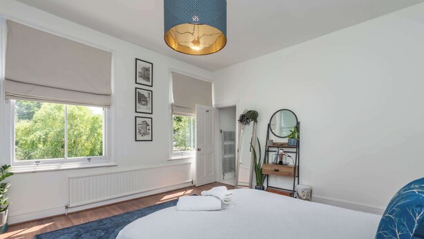 Une chambre avec un grand lit, une fenêtre avec des stores, un luminaire au plafond et une étagère fixée au mur, agrémentée d’un miroir et d’objets décoratifs.