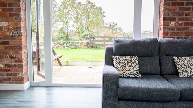 Een grijze bank met gekleurde kussens in een kamer met een bakstenen muur en een schuifdeur die leidt naar een terras met een houten omheining en een gazon.