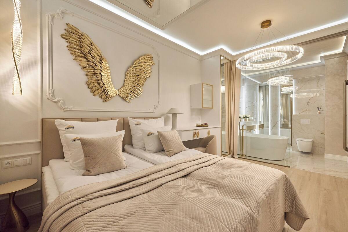 Un dormitorio con una cama grande, un adorno mural con forma de alas, una araña y un baño con ducha de vidrio.