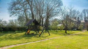 A playground with a swing set and climbing structure in a grassy area with trees and a house in the background. - Y Wennol - sleeps 8 guests  in 3 bedrooms (Llangefni)