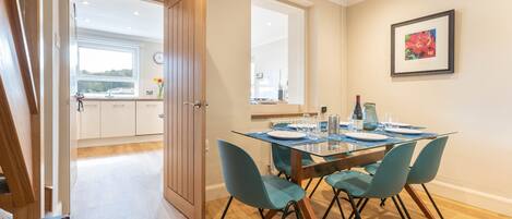 Un coin repas avec une table en bois dressée pour quatre personnes, des chaises de couleur turquoise et une vue sur la cuisine par une porte ouverte.