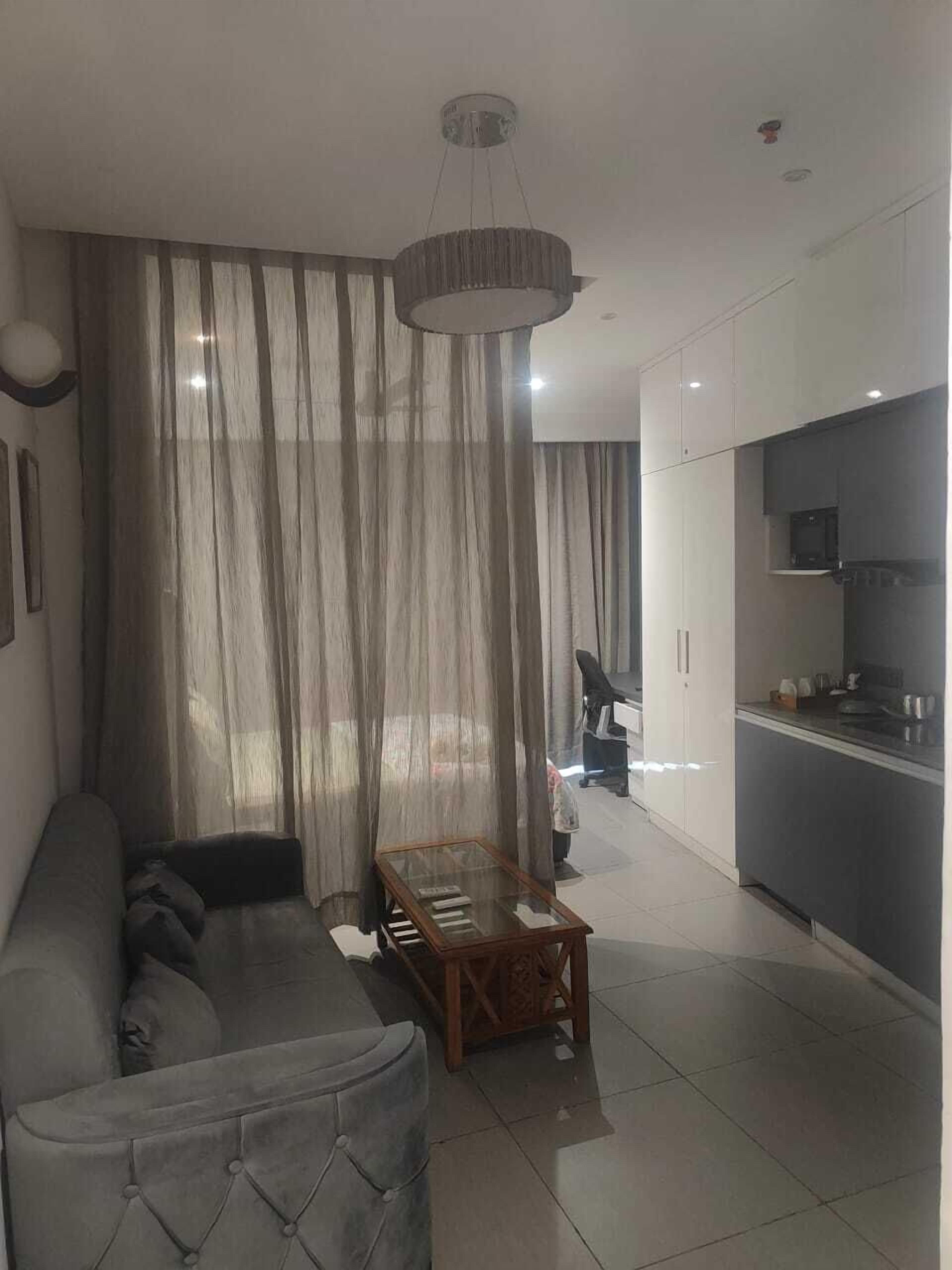 A modern living room with a grey sofa, a glass coffee table, and a kitchen area with a grey countertop and cabinets.