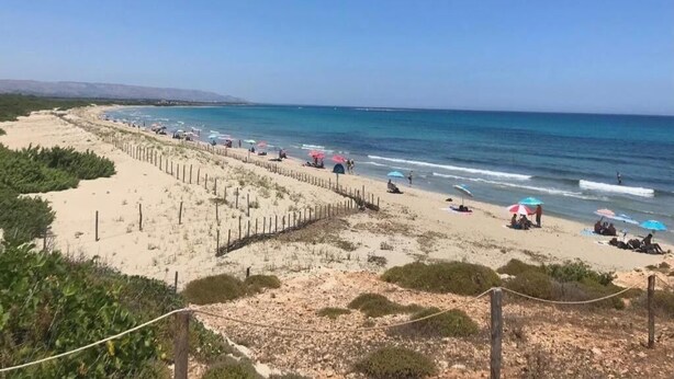A beach with a fence, umbrellas, and people.