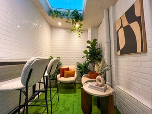 A cozy corner with a white brick wall, a green artificial turf floor, a small round table with books, and a chair with a white cushion. - UrbanFlat 11 - Cosy king studio near champs Elysees (París)