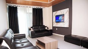 A modern living room with a black leather sofa, a wooden coffee table, and a black ottoman. A flat-screen TV is mounted on the wall, and the room has a neutral color palette. - The Moon Crib (Gisenyi)