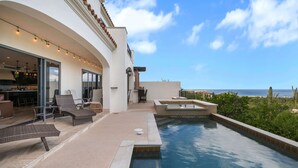 A modern outdoor pool area with a view of the ocean, a patio with seating, and a kitchen visible through glass doors. - Villa Azulejos, Luxury villa with privacy and breathtaking views (Cabo San Lucas)