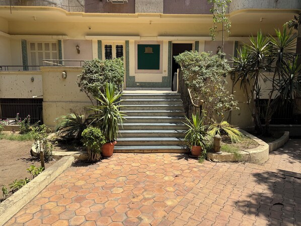 A building entrance with a staircase leading to a door, surrounded by potted plants and a tiled pathway.