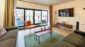 Ein modernes Wohnzimmer mit Sofa, Couchtisch, Fernseher und Blick auf die Straße durch große Fenster.