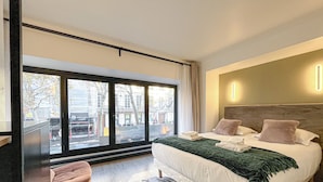 Ein modernes Hotelzimmer mit großem Fenster, einem Bett mit Kissen, einem Nachttisch und Blick auf die Straße.