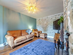 Una sala de estar con un sofá, un sillón más pequeño, un tapete azul, una pared de piedra y una chimenea con una planta.