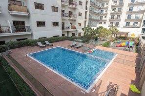 Piscina no terraço com espreguiçadeiras, rodeada por um jardim e um edifício com várias andares e varandas.