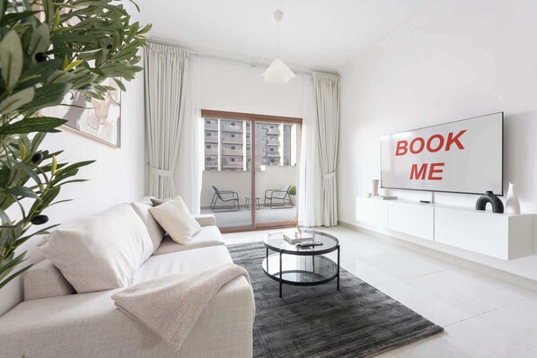 Uma sala de estar moderna com um sofá, uma mesa de centro, uma TV exibindo o texto 'BOOK ME' e uma varanda com vista para prédios.