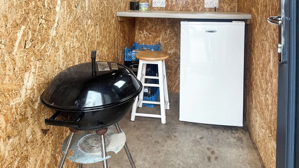 A compact kitchen with a black grill, a white refrigerator, and a stool.